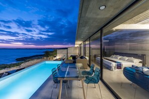 Uma área externa moderna com mesa e cadeiras de vidro, piscina e vista para o oceano ao entardecer.