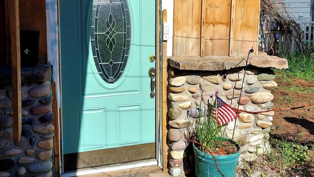 A turquoise door with a decorative glass panel, a stone wall with a wooden door, and a small garden with a potted plant and an American flag. - Snug and Cozy Hideaway is close to National Parks, BYU Idaho, and River Beach (St Anthony)
