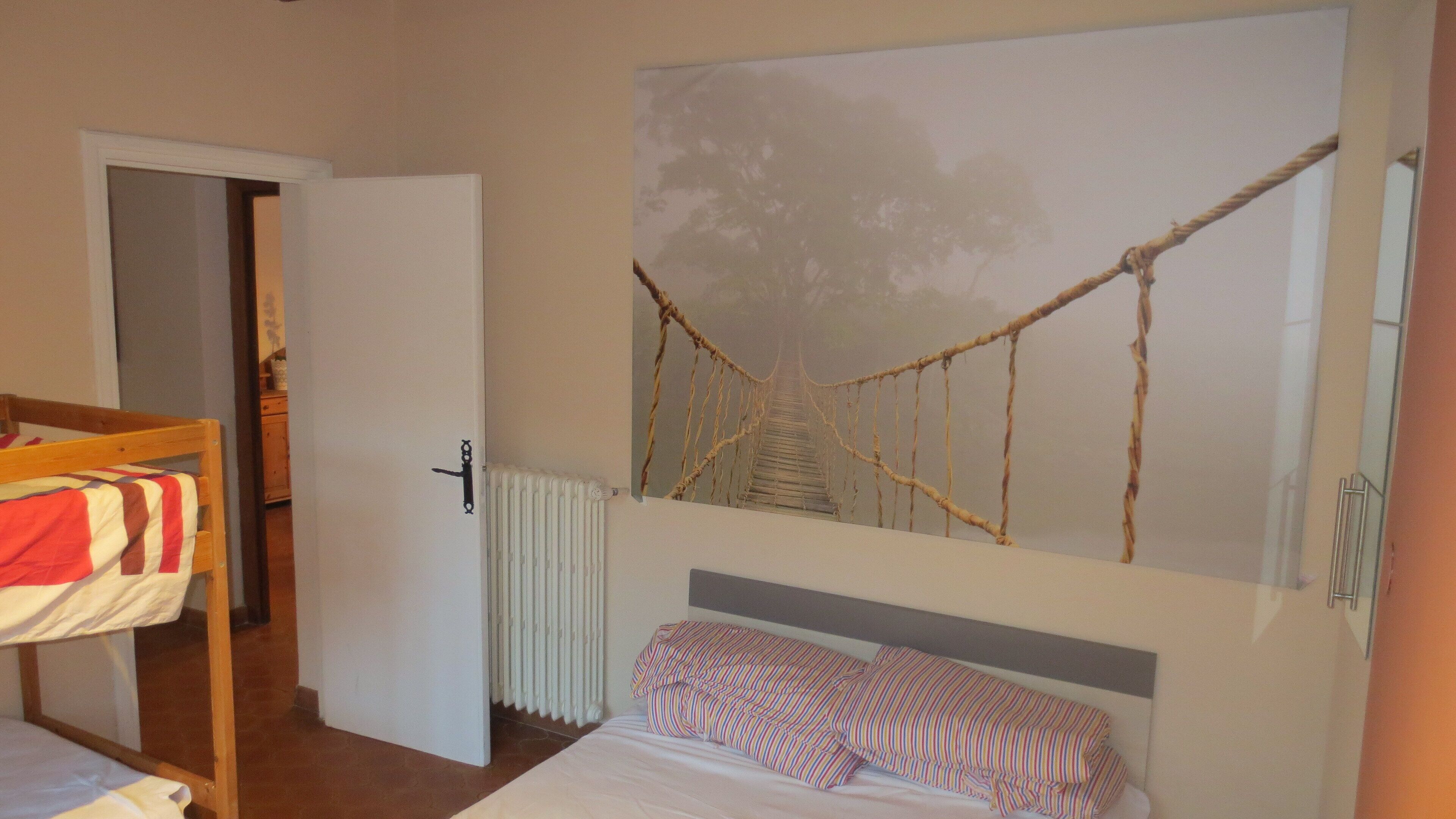 Un dormitorio con literas, un radiador, una obra de arte colgada en la pared que representa un puente colgante y una puerta que da a otra habitación.