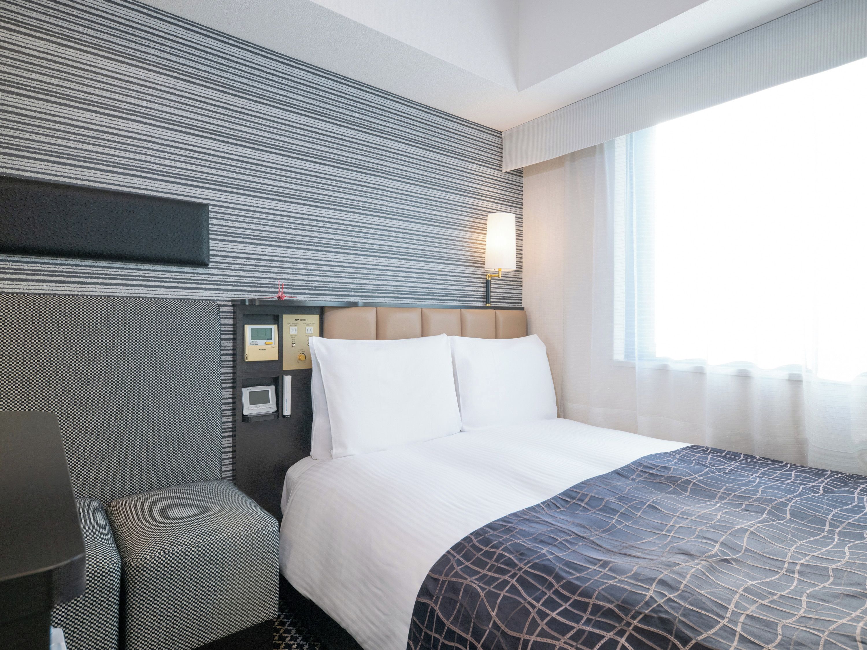 Une chambre d’hôtel équipée d’un lit, d’un bureau, d’une chaise, d’un panneau de commande et d’une fenêtre avec des rideaux.