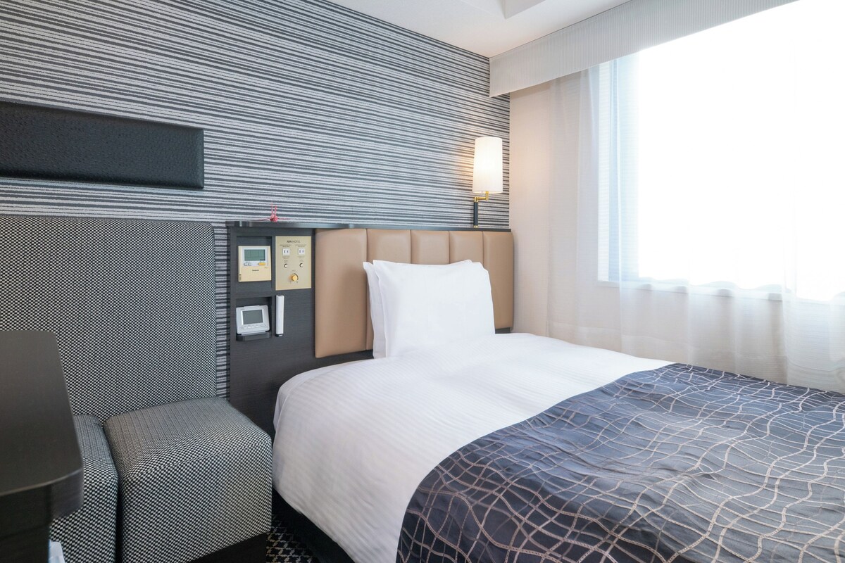 A hotel room with a bed, a bedside table, a control panel, and a window with curtains.