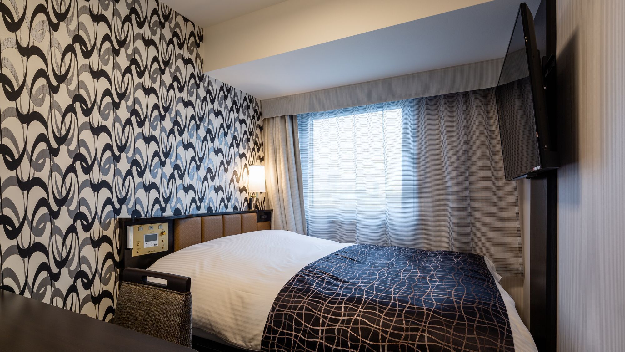 A hotel room with a patterned wallpaper, a bed with a geometric-patterned bedspread, a bedside table with a lamp, and a television mounted on the wall.
