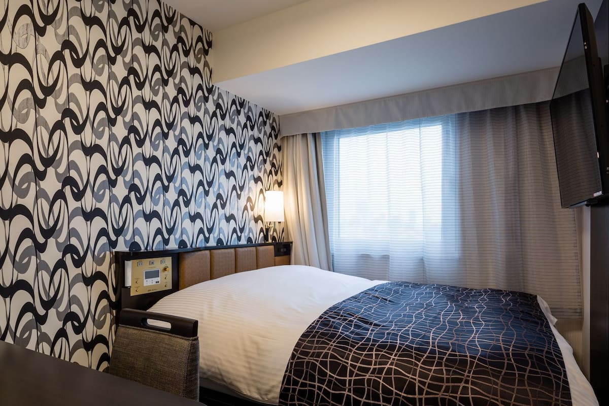 A hotel room with a patterned wallpaper, a bed with a geometric-patterned bedspread, a bedside table with a lamp, and a television mounted on the wall.
