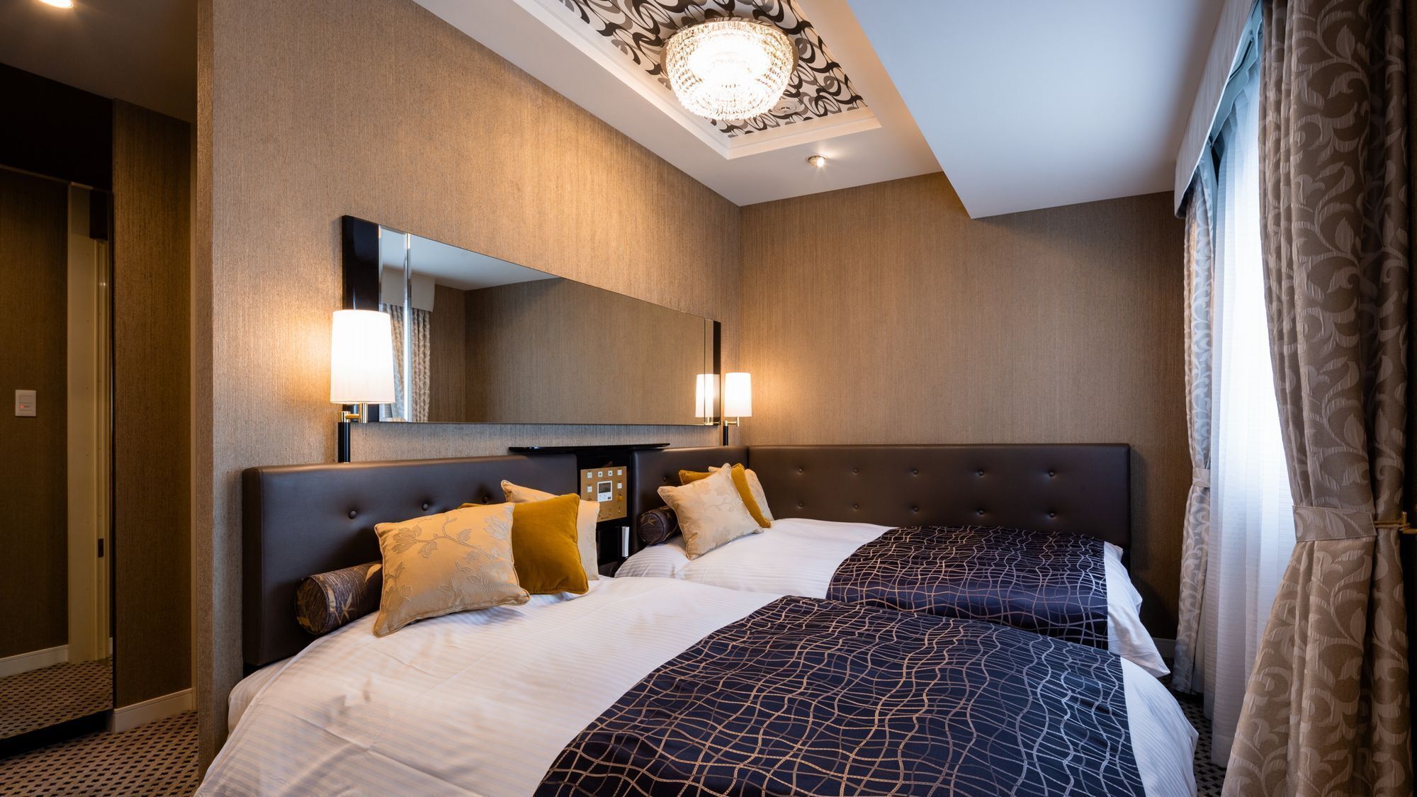 A hotel room with two beds, a large mirror, wall sconces, and a chandelier.