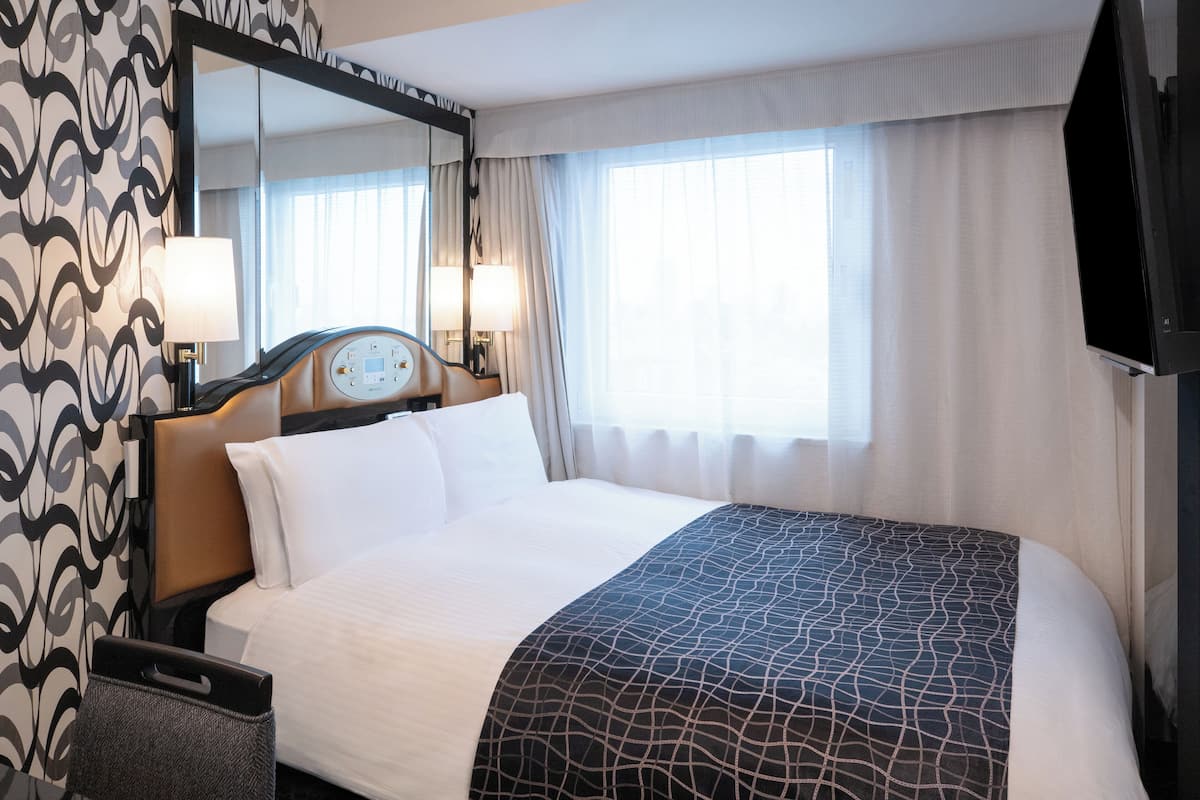 A hotel room with a large bed, a bedside table with a lamp, a mirror, and a window with curtains.