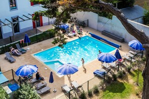 Ein Swimmingpool mit Liegestühlen und Sonnenschirmen, umgeben von einem eingezäunten Bereich mit einem Gebäude im Hintergrund.
