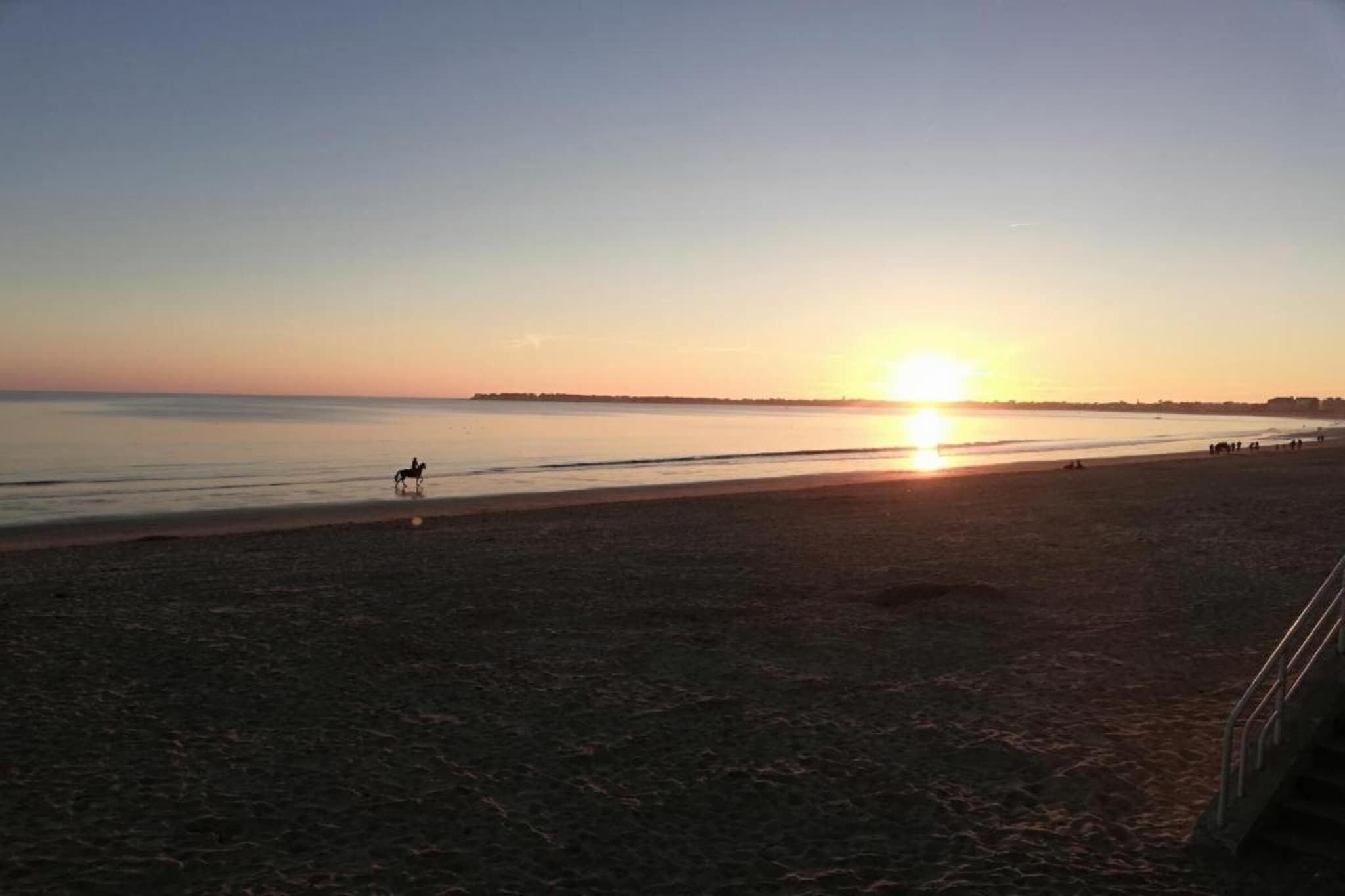 Una playa al atardecer con un caballo y jinete, mar tranquilo y costa lejana.