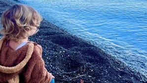 Un bambino seduto su una riva ciottolosa che si affaccia su un lago tranquillo, con barche e uno sfondo boschivo.