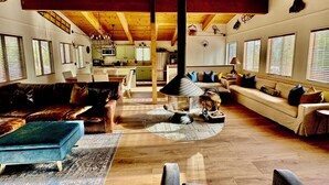 Un'ampia zona giorno con soffitto alto, travi in legno, un camino al centro e una cucina sullo sfondo.