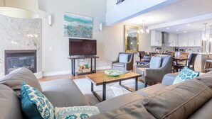 A spacious living room with a fireplace, a television, and a dining area with a kitchen in the background.
