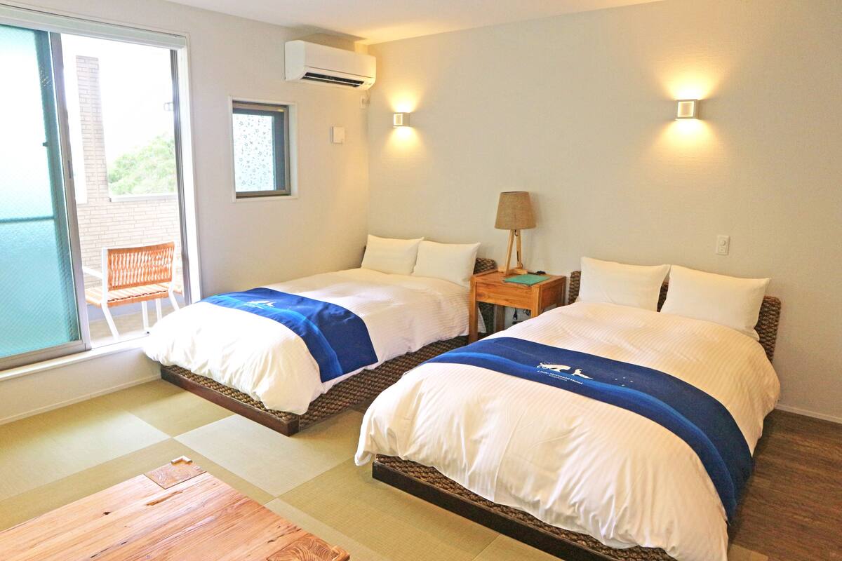 A hotel room with two beds, a sliding glass door to the balcony, and a small table with a lamp.