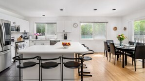 A modern kitchen with a central island, bar stools, a dining area with a table and chairs, and large windows providing natural light. - Hamptons Retreat w/ Pool & Tennis - 90min from NYC (Quogue)
