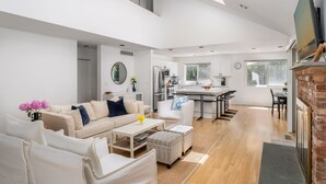 A spacious living area with a sofa, coffee table, and ottomans, a kitchen with a dining area, and a fireplace. - Hamptons Retreat w/ Pool & Tennis - 90min from NYC (Quogue)