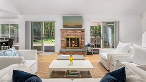 A modern living room with a fireplace, white sofas, a coffee table, and large glass doors leading to an outdoor area. - Hamptons Retreat w/ Pool & Tennis - 90min from NYC (Quogue)