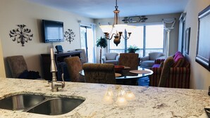 A kitchen with a granite countertop, stainless steel sink, and a dishwasher. There is a dining area with a round table and chairs, and a living room with a sofa and a TV mounted on the wall.