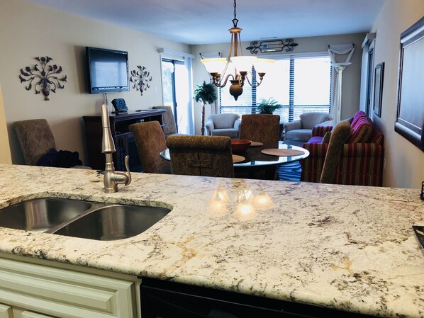 A kitchen with a granite countertop, stainless steel sink, and a dishwasher. There is a dining area with a round table and chairs, and a living room with a sofa and a TV mounted on the wall.