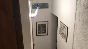 A narrow hallway with a wooden door on the left, a white textured wall on the right, and two framed artworks on the wall. - Apartment in the historic center (Ferrara)