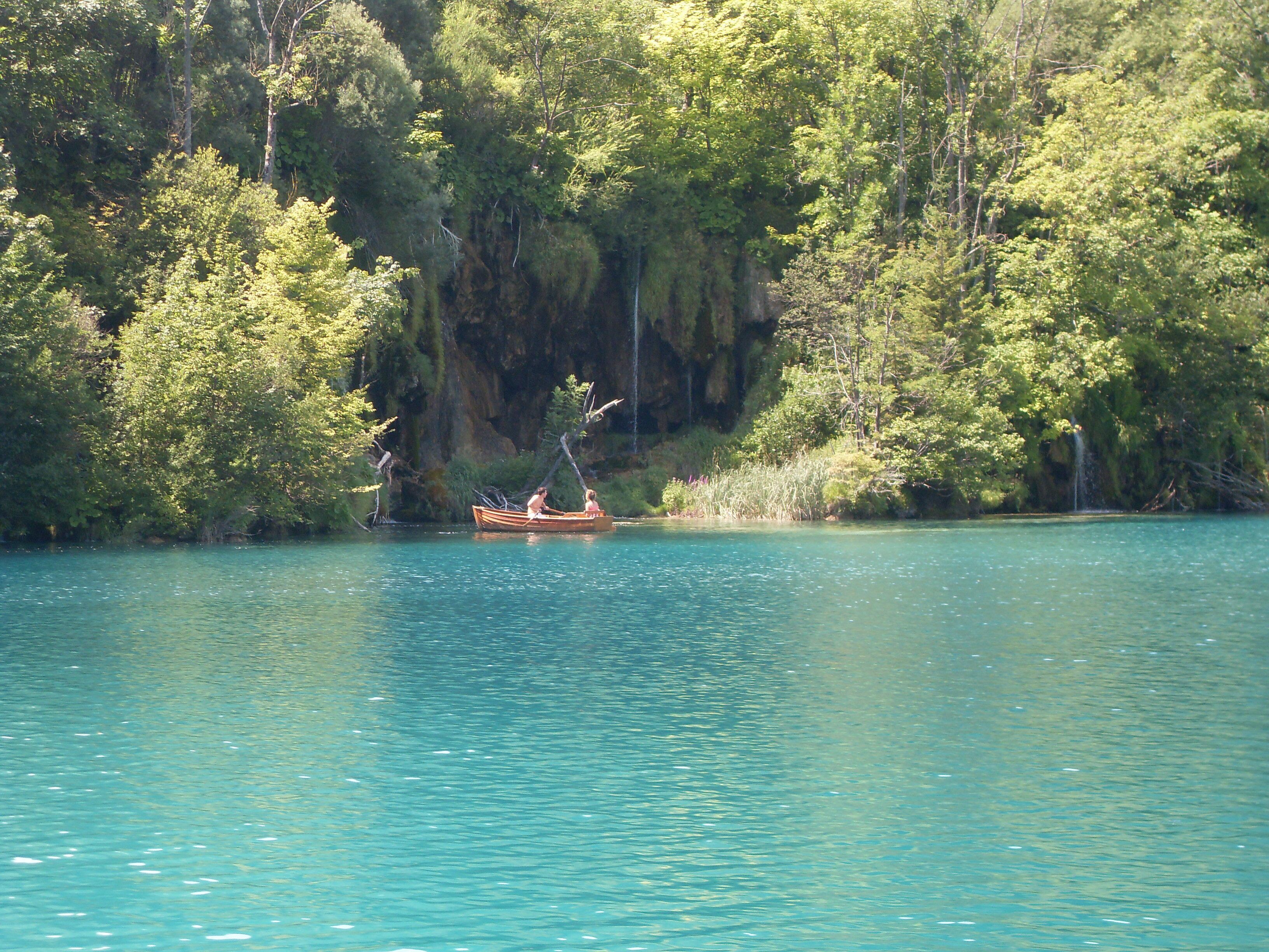 A calm lake with a wooden boat carrying two people, surrounded by dense forest and rocky cliffs.