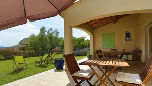 Une terrasse extérieure avec une table et des chaises, un espace abrité et une vue sur un jardin et une maison en arrière-plan.