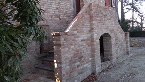 A rustic stone building with a brick staircase leading to a door, surrounded by trees and a gravel path.