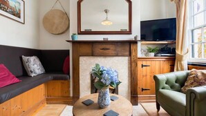 Uma sala de estar acolhedora com um sofá, uma poltrona verde, uma mesa de centro de madeira com um vaso de flores, uma lareira e uma televisão.