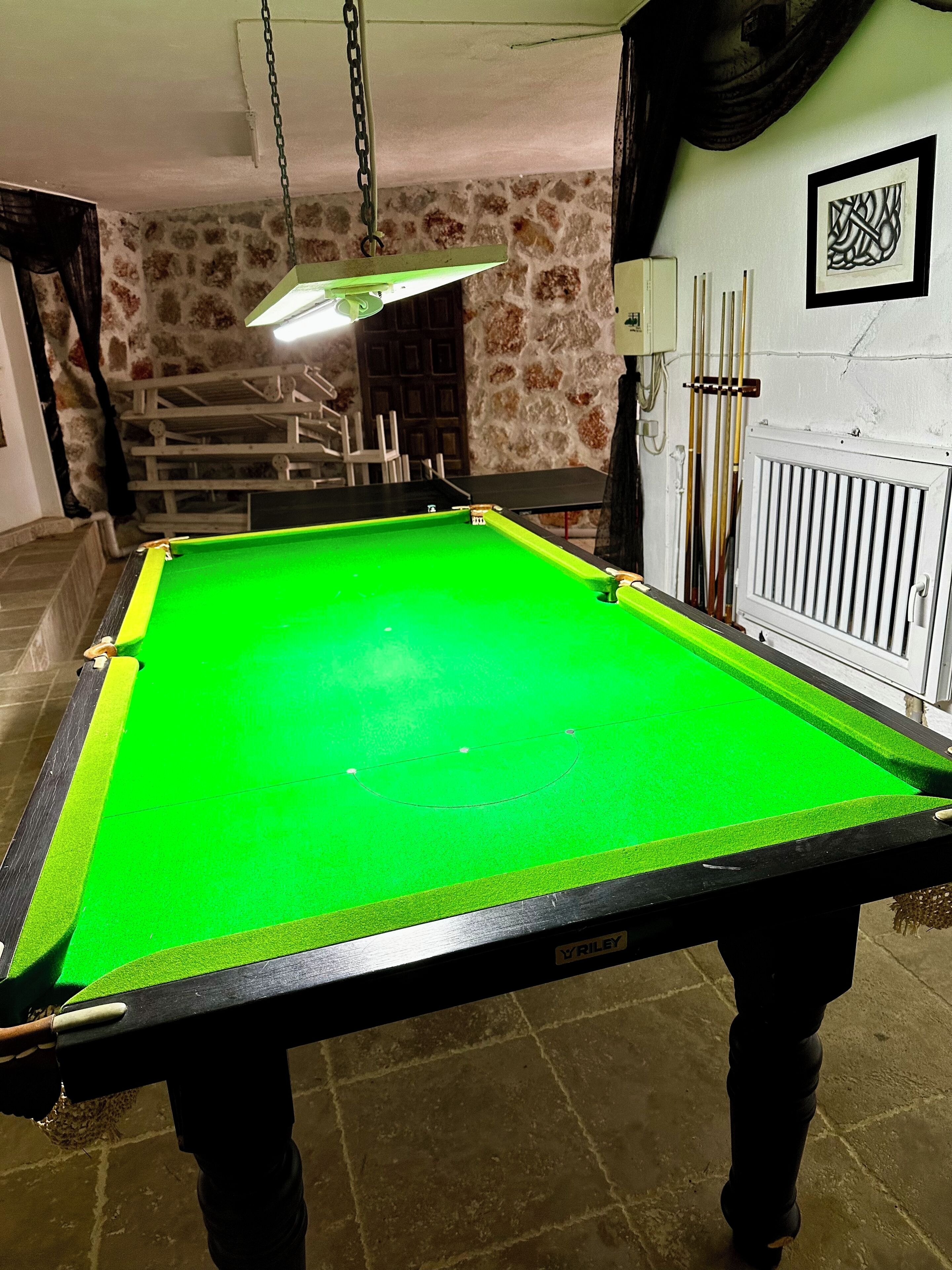 Een pooltafel met een groene viltbekleding, een hangende lamp en een stenen muur op de achtergrond.