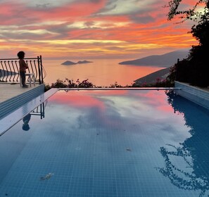 An infinity pool with a view of a sunset over water and hills.