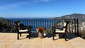 Un balcón con dos sillas de madera y una mesita pequeña, con vistas a la costa y un cielo despejado.