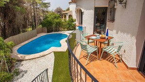 Un área de piscina con solárium empedrado, una mesa con sillas y vistas a los árboles y edificios circundantes.