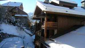 A wooden chalet with a balcony, surrounded by snow-covered terrain and other buildings.