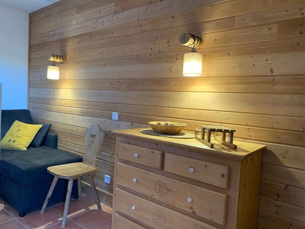 A cozy room with a wooden wall, a blue sofa with a yellow pillow, a wooden chair, and a wooden dresser with drawers.