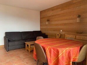 A room with a sofa, a small table, a dining table with a tablecloth, and wooden panel walls.