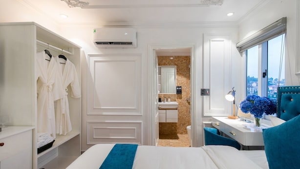 A hotel room with a bed, a wardrobe, a vanity with a mirror, a desk with a lamp, and a window with a city view. - Diamond Hotel Sapa (Sa Pa)