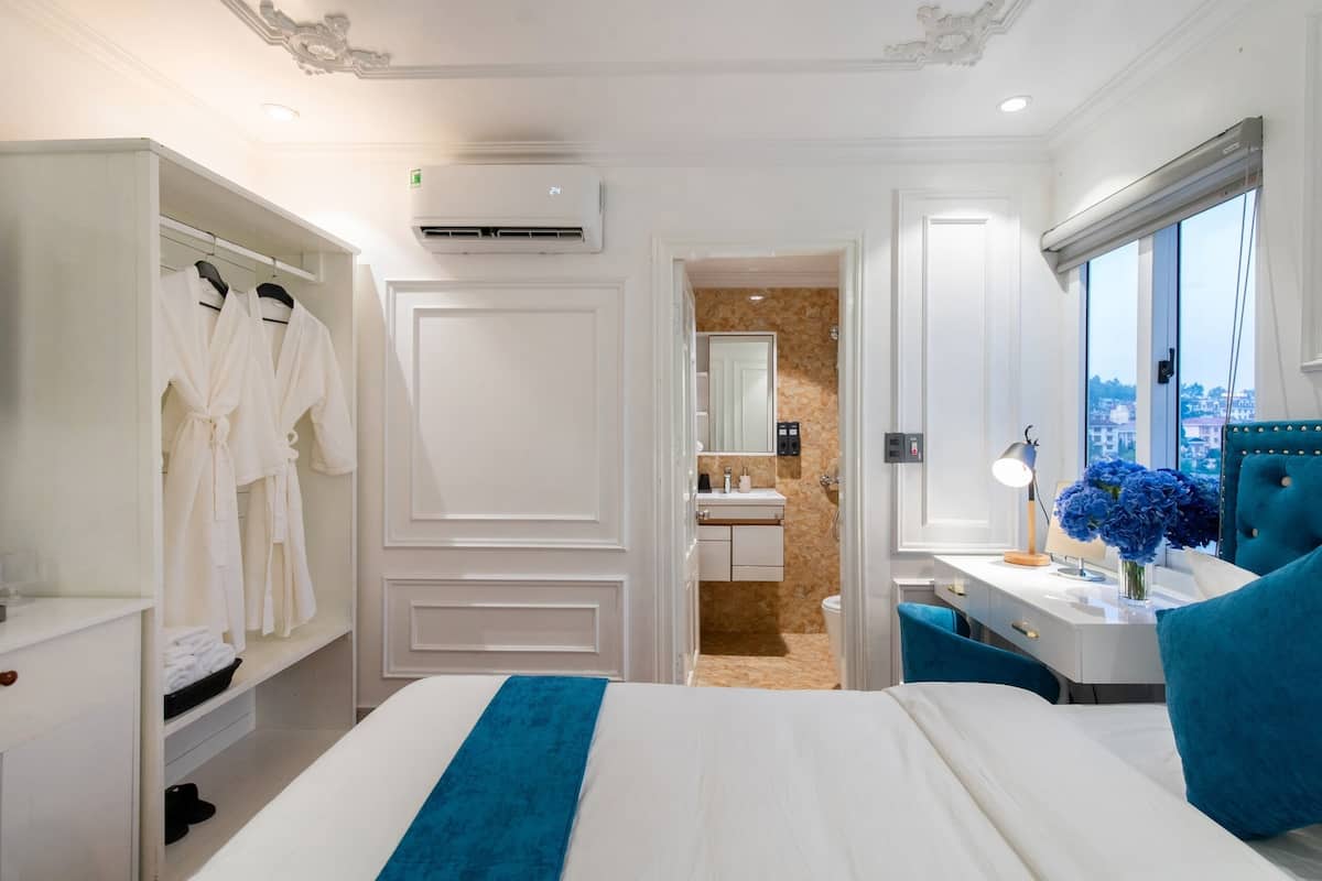 A hotel room with a bed, a wardrobe, a vanity with a mirror, a desk with a lamp, and a window with a city view.