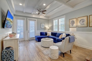 A spacious living room with a blue sectional sofa, a ceiling fan, and a large TV on the wall.