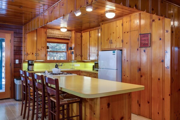 A cabin-style kitchen with wooden walls, a central island with bar stools, a refrigerator, and a window above the sink.