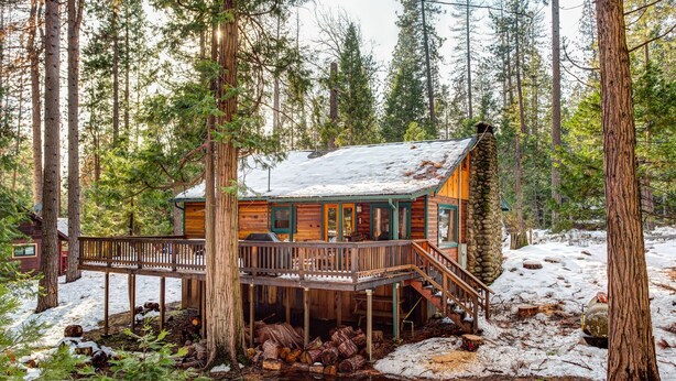 A cabin with a wooden deck and a stone foundation, surrounded by tall trees and snow. - (59) Hansen Cabin at The Redwoods In Yosemite (Yosemite National Park)