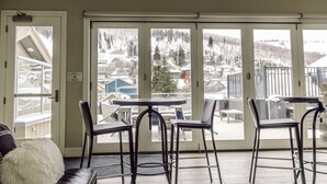 A room with a large sliding glass door leading to a balcony with a view of a snowy mountain and houses. There are four black chairs and a small round table on the balcony. -   Luxury Ski-In/Out Town Lift Walk to Main St (Park City)