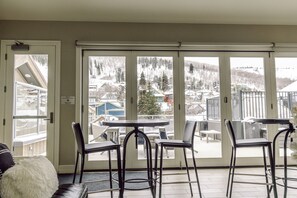 A room with a large sliding glass door leading to a balcony with a view of a snowy mountain and houses. There are four black chairs and a small round table on the balcony.