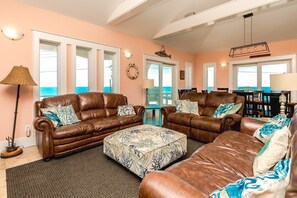 A living room with leather sofas, a patterned ottoman, and a view of the ocean through large windows. - 99 Oleander - Luxury 9 BR Home - Great Views, Pool, Hot Tub, Elevator & More! (Surf City)