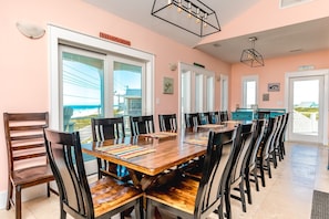 A dining area with a long wooden table and chairs, large windows with a view of the beach, and a sign that reads 'BEACH BUMS WELCOME'. - 99 Oleander - Luxury 9 BR Home - Great Views, Pool, Hot Tub, Elevator & More! (Surf City)