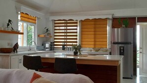 Una cocina moderna con una isla central, persianas de madera, un refrigerador de acero inoxidable y un comedor con una silla.