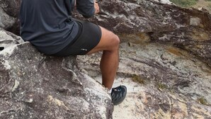 Una persona sentada sobre un saliente rocoso, con vistas a un paisaje costero y colinas distantes.