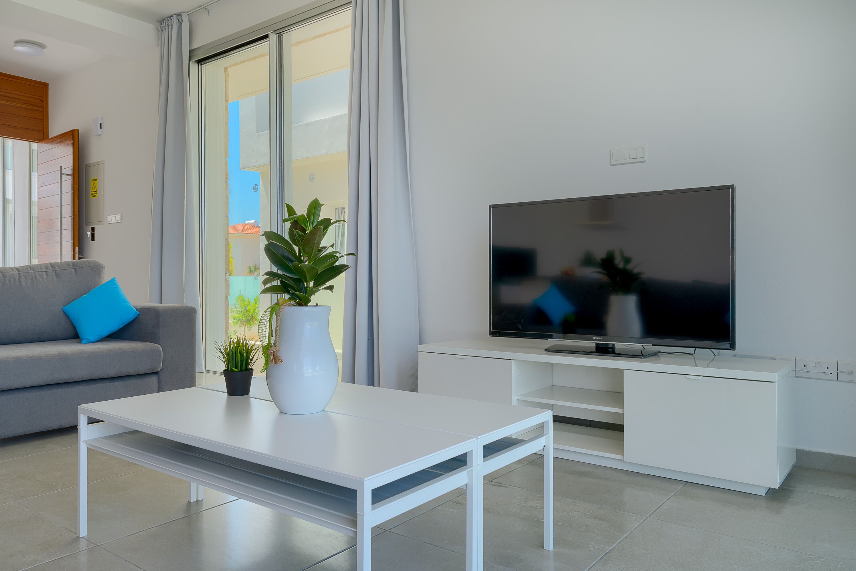 A modern living room with a grey sofa, a white coffee table, a TV on a white stand, and a sliding glass door leading to a balcony.
