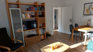 A living room with a wooden bookshelf, a TV, a coffee table, and a dining area with chairs and a table. - Stylish apartment in a fantastic location right on Volkspark (FL)