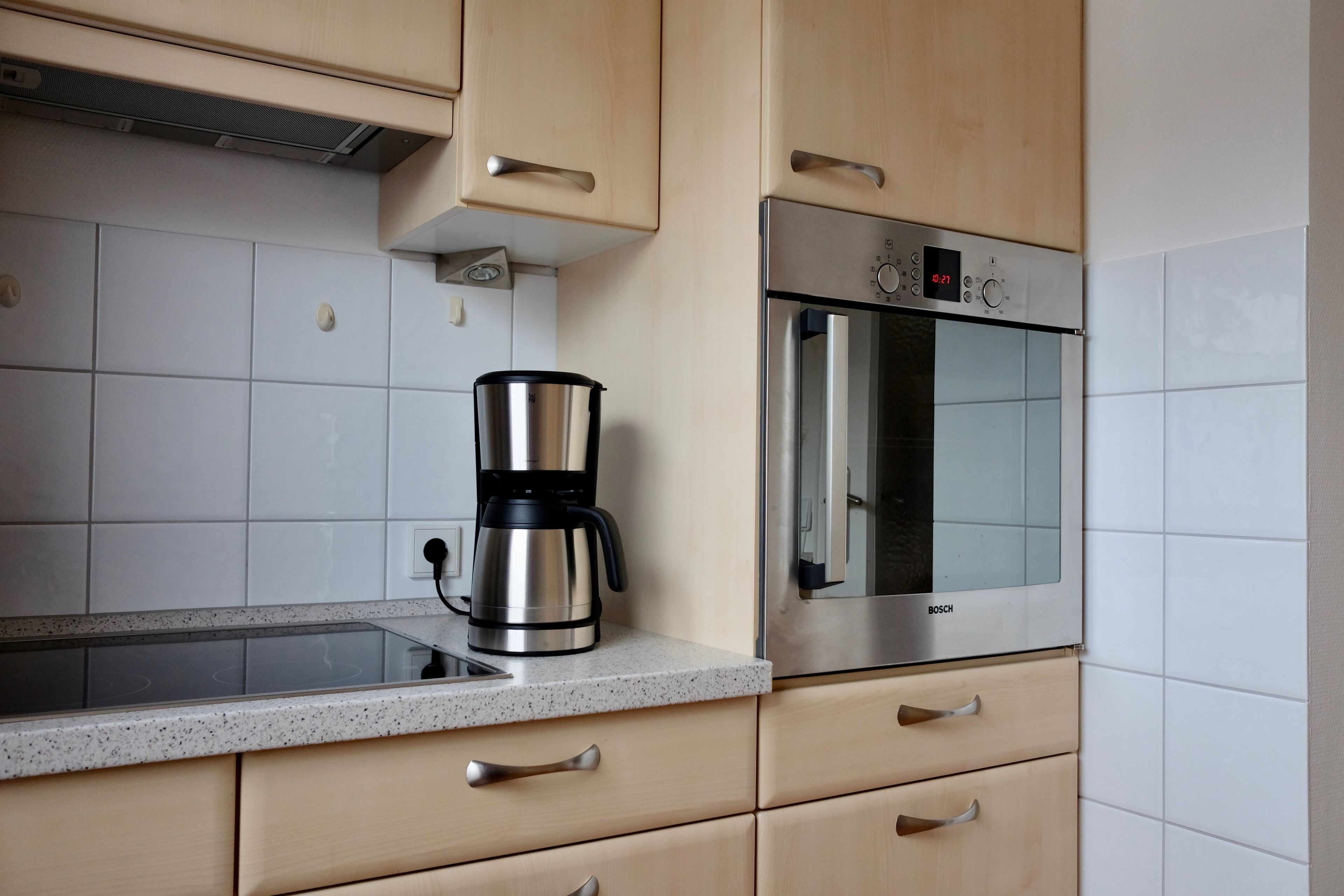 A kitchen with a stainless steel coffee maker, a Bosch oven, and white tiled walls.
