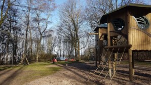 A wooden play structure with a slide and climbing elements, surrounded by trees and a dirt path. - Stylish apartment in a fantastic location right on Volkspark (FL)