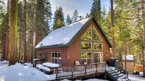Una cabaña con fachada de madera, ventanas grandes, una terraza y un techo cubierto de nieve.