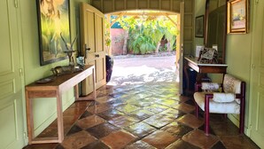 Un pasillo con piso de baldosas, un banco de madera, un cuadro enmarcado en la pared y una puerta que da a un área exterior con plantas.