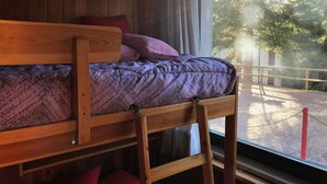 A wooden bunk bed with a patterned blanket and pillows, a window with a view of trees and a fence outside.